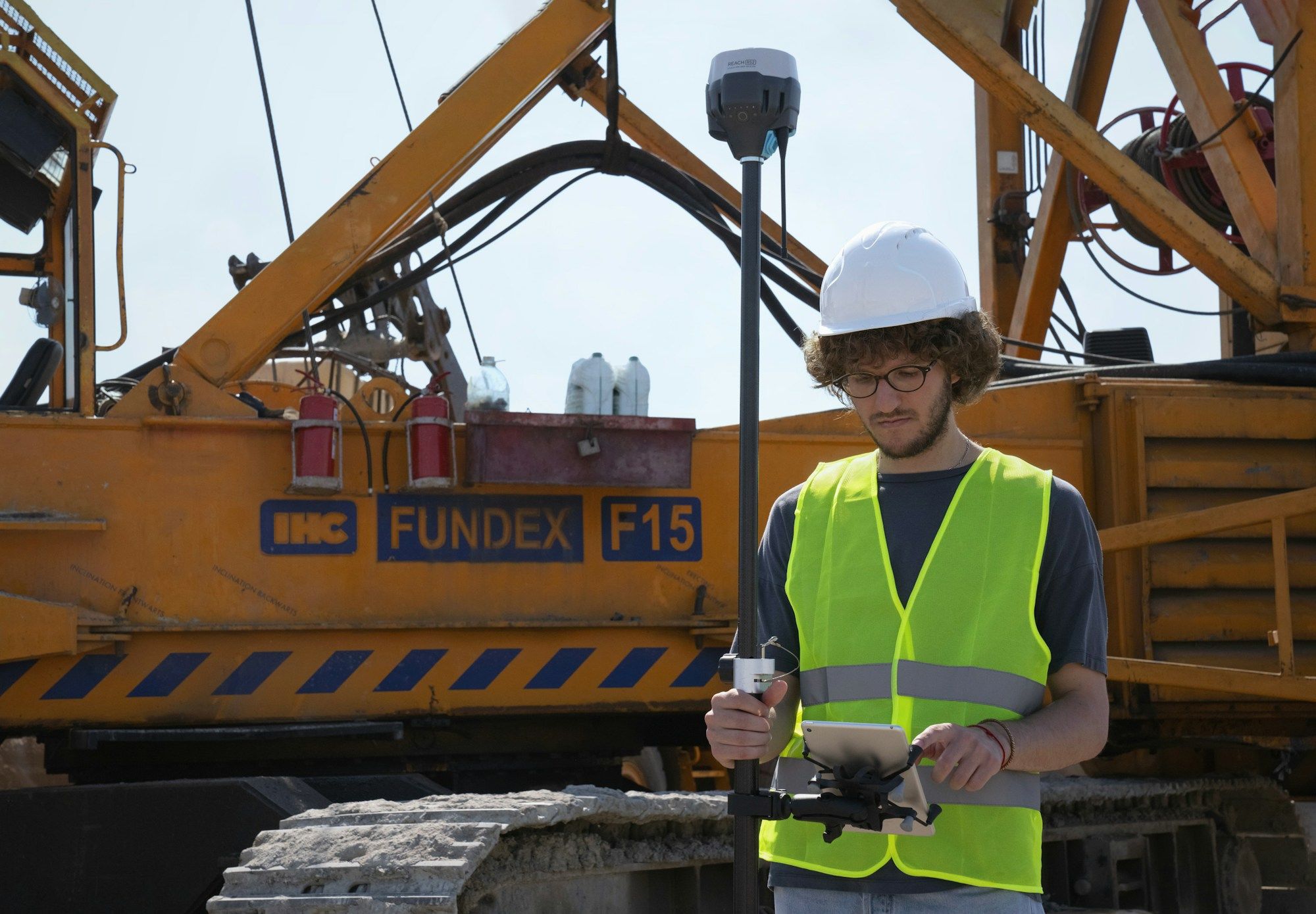Worker in a hard hat and high-visibility vest checking a device beside heavy plant
